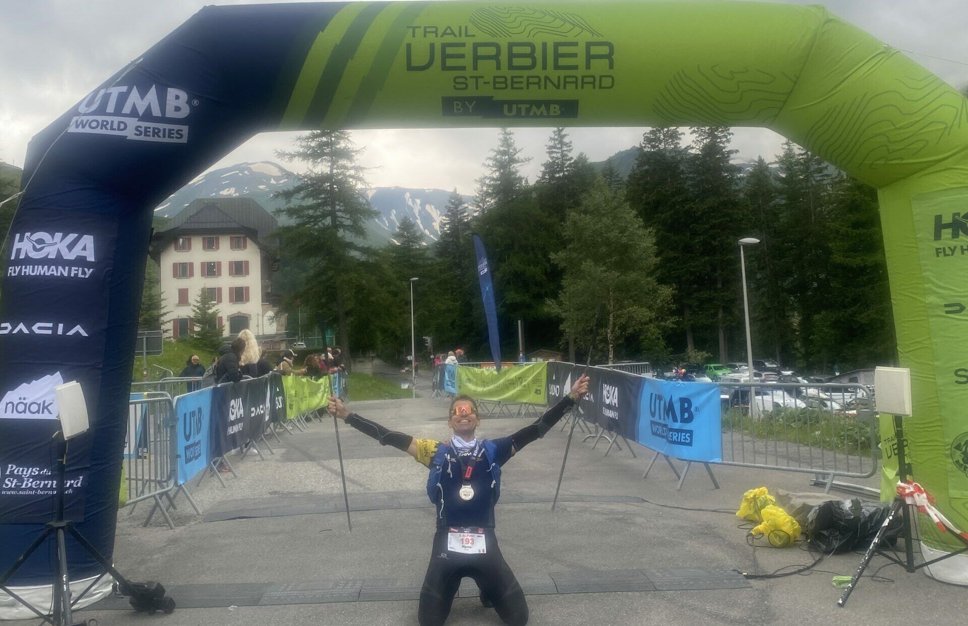 UTMB Verbier arrivo TRM Team MArco Mori inginocchiato sotto il gonfalone dellarrivo scaled e1725980552426 ad43b765